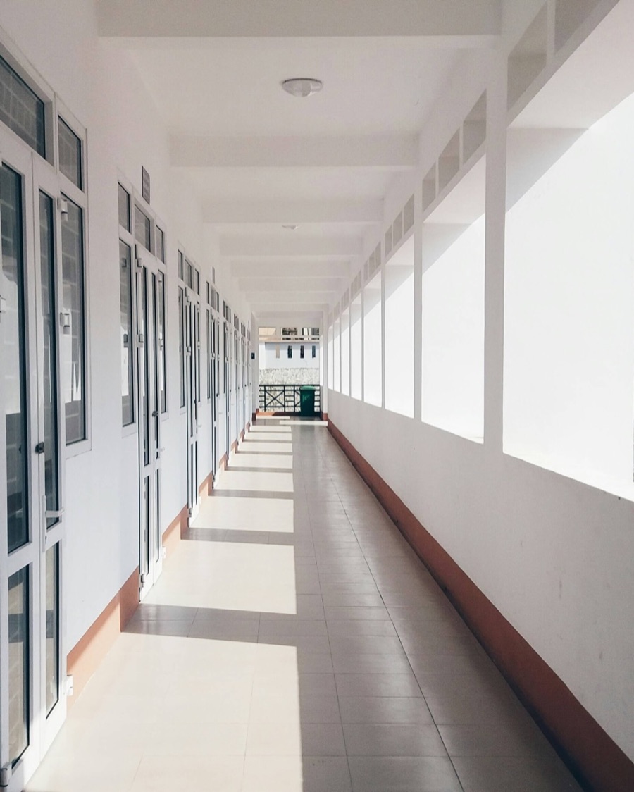 A sunlit school corridor, glazed classroom doors along one side and open windows along the other, tiled floor receding into the distance