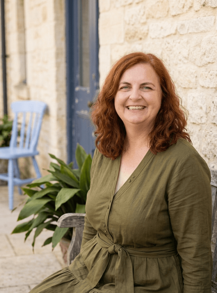 Ev Abbott, leadership coach, smiling and seated on a wooden bench outdoors, wearing an olive-green dress, beside a leafy plant and a soft-blue chair against a honey-coloured stone wall